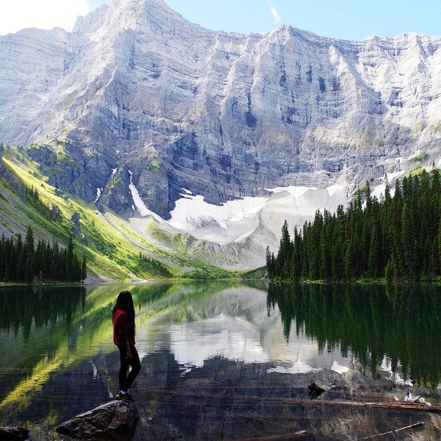 A woman stands on the shore of Rawson Lake during the summertime, looking up at the overarching Mt. Sarrail.